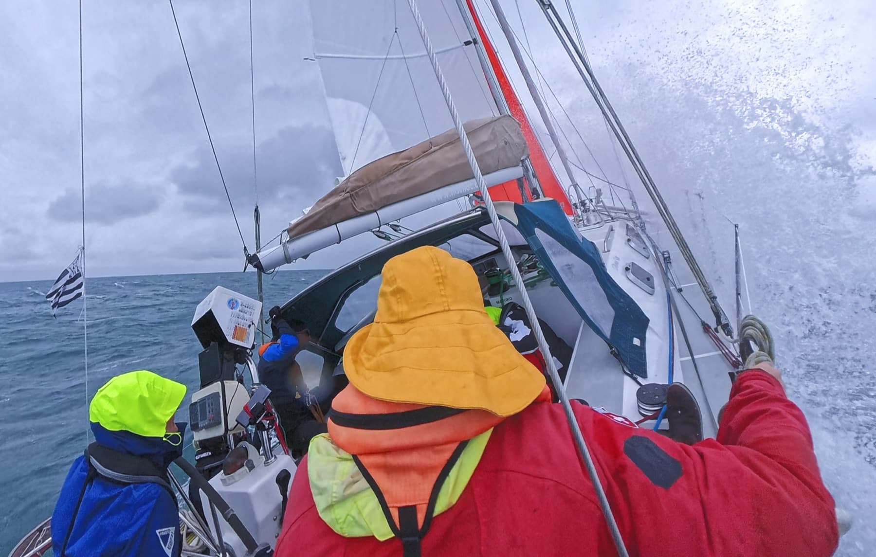 Crew im Regenzeug an Deck bei Spritzwasser — die echte Bretagne-Praxis.