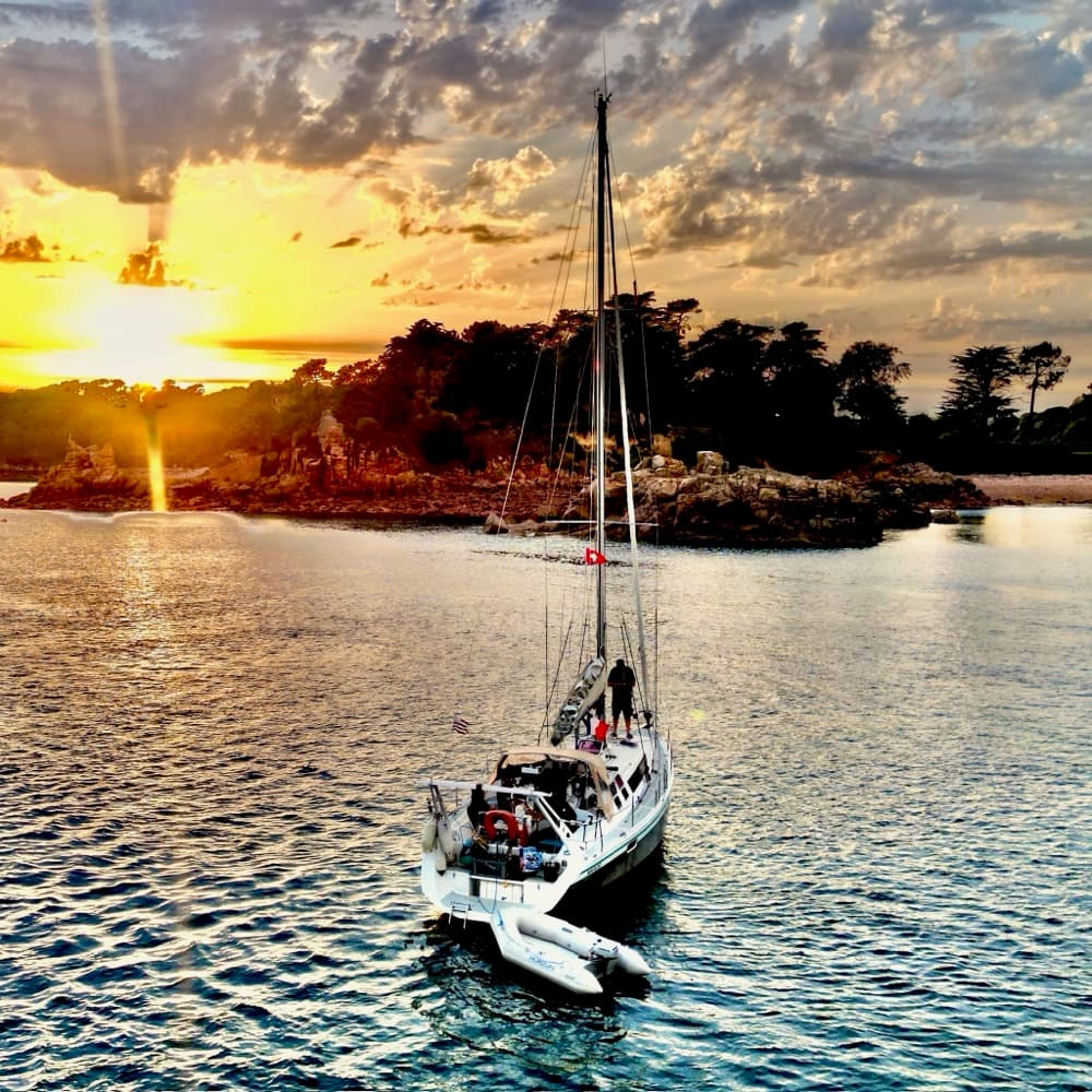 Segelyacht mit Beiboot vor Anker im Sonnenuntergang, Crew an Deck vor einer bewaldeten Felsinsel