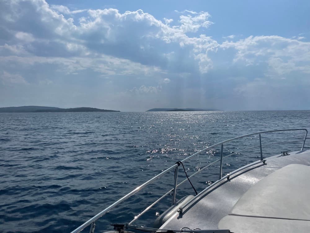 Blick über den Bug aufs offene Meer, Sonnenstrahlen brechen durch die Wolken über den Inseln
