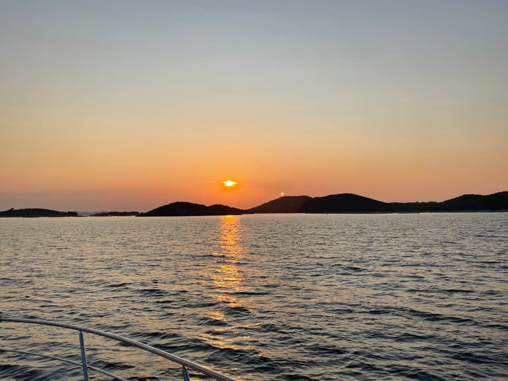 Sonnenuntergang über einer Inselkette, die Sonne spiegelt sich im Wasser – Blick von Bord