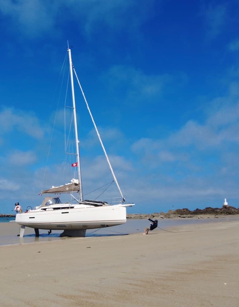 Trockengefallene Segelyacht mit Schweizer Flagge am Strand, im Hintergrund ein kleiner Leuchtturm